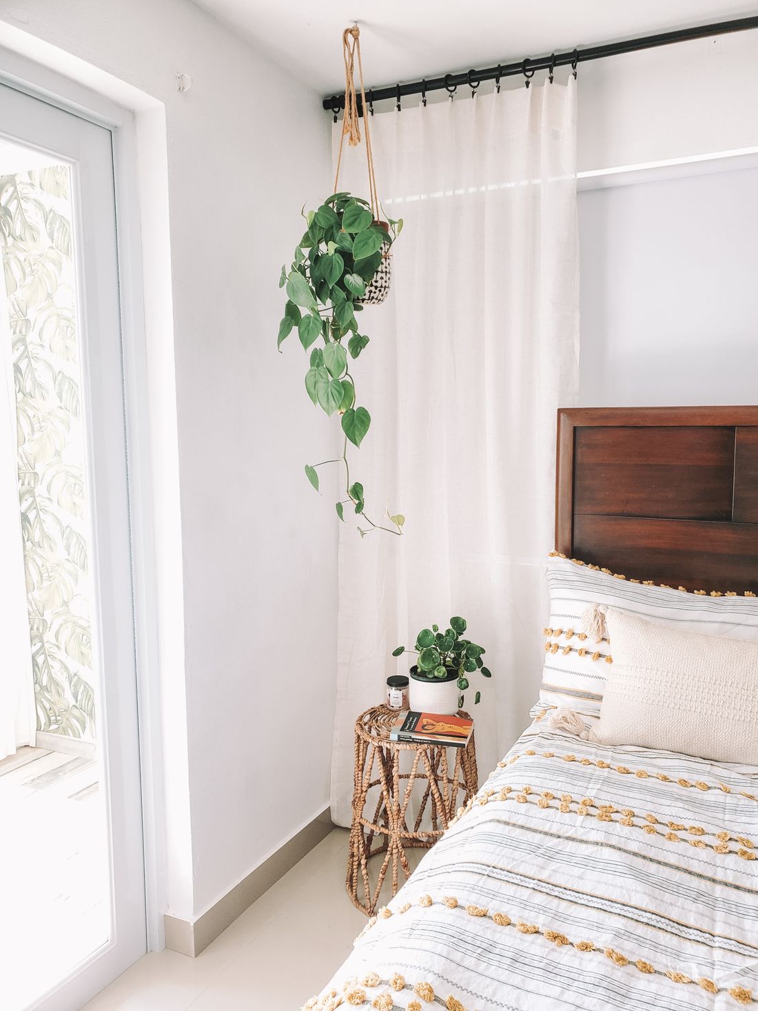 A bedroom with a trailing philodendron hanging from the ceiling beside a curtained door, and a small pilea on a side table by the bed