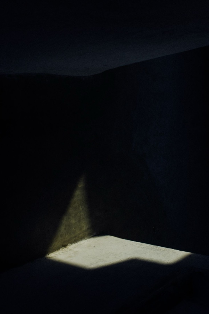 The corner of a dark, empty room with a single small patch of daylight lying on the floor and the walls falling away into black