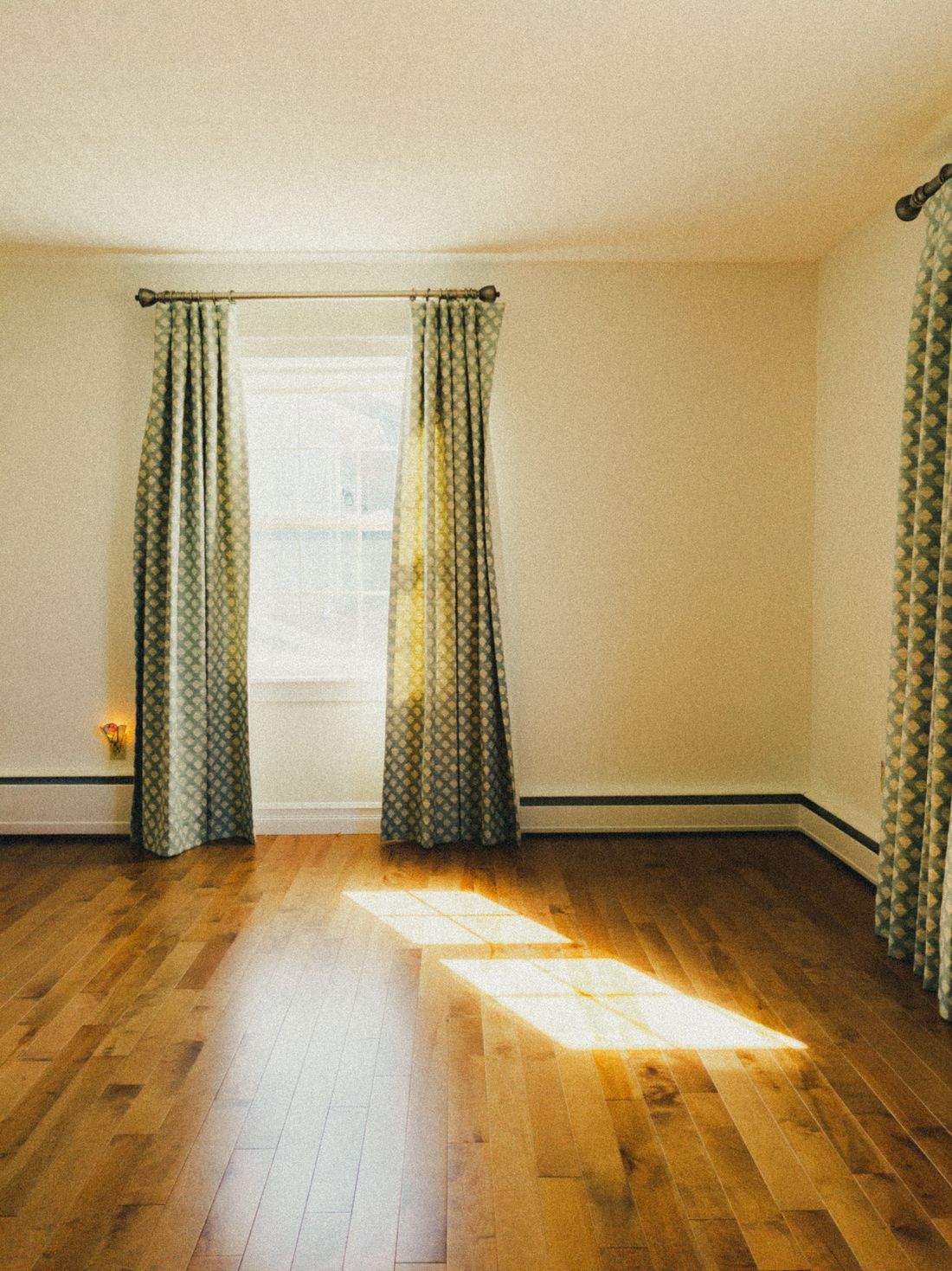 A curtained window in an empty room with one bar of sunlight lying across the floorboards