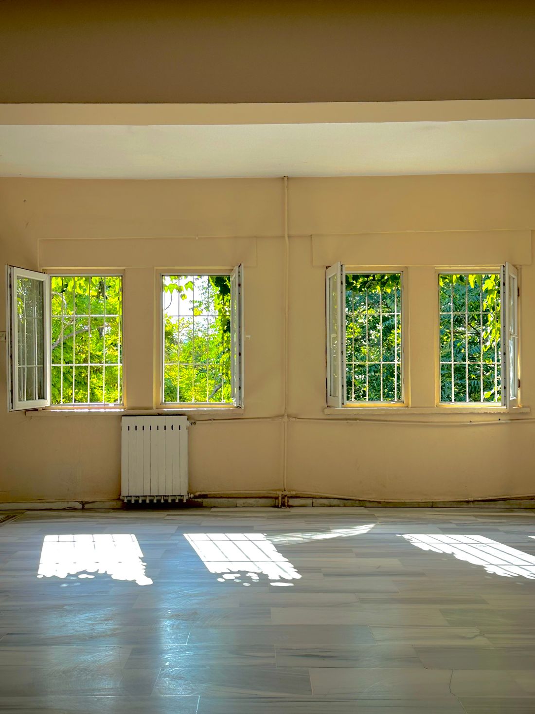 An empty room with four casement windows along one wall, sunlight throwing their grids onto a marble floor several metres in