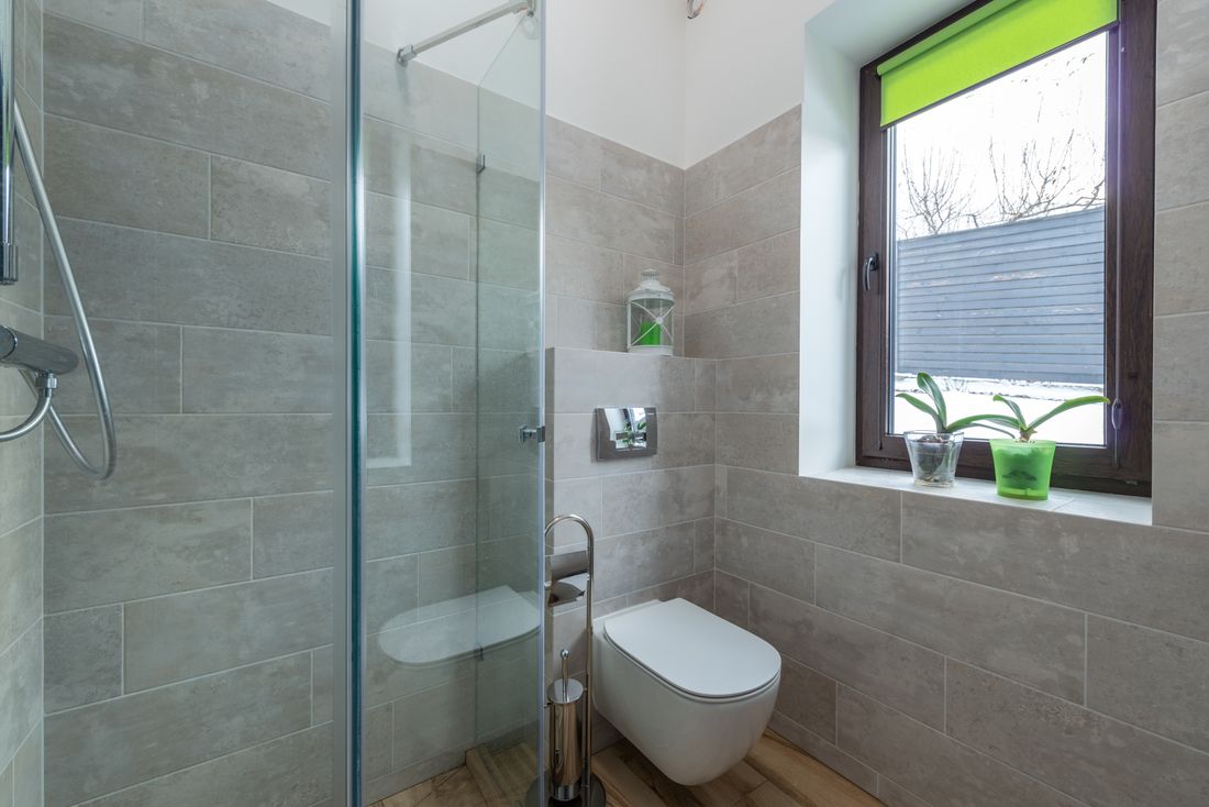 A tiled bathroom with a glass shower enclosure and a small window on the far wall, two orchids on the sill